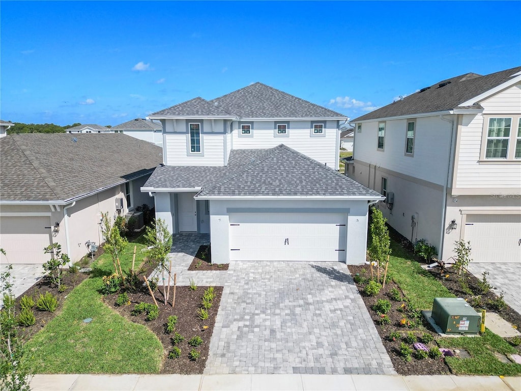 traditional-style house with decorative driveway, an attached garage, central air condition unit, stucco siding, and a shingled roof