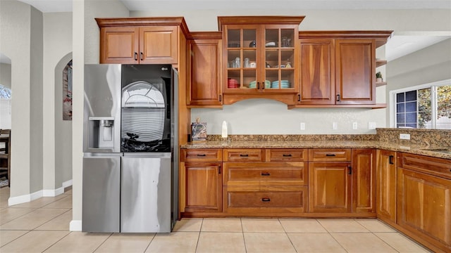kitchen featuring stainless steel fridge with ice dispenser, stone counters, brown cabinets, light tile patterned floors, and glass insert cabinets