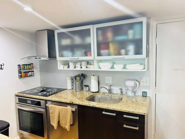 kitchen with light stone countertops, glass insert cabinets, a sink, wall chimney range hood, and stainless steel appliances