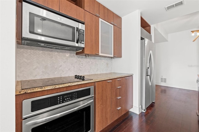 kitchen with appliances with stainless steel finishes, brown cabinetry, decorative backsplash, dark wood-type flooring, and light stone counters