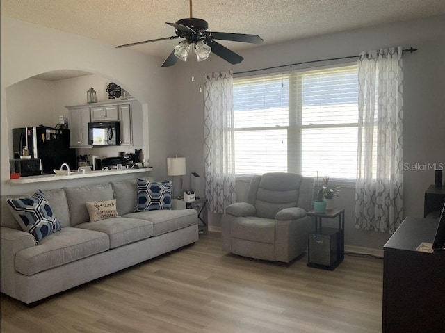 living room featuring light wood finished floors, a textured ceiling, arched walkways, a ceiling fan, and baseboards