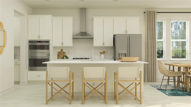 kitchen with refrigerator with ice dispenser, a sink, wall chimney range hood, double oven, and an island with sink