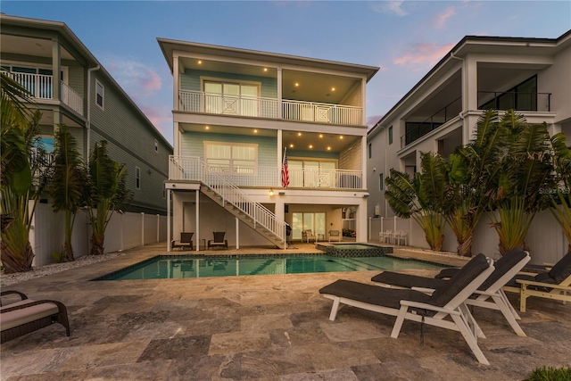 back of house at dusk featuring a balcony, stairway, a fenced backyard, a pool with connected hot tub, and a patio