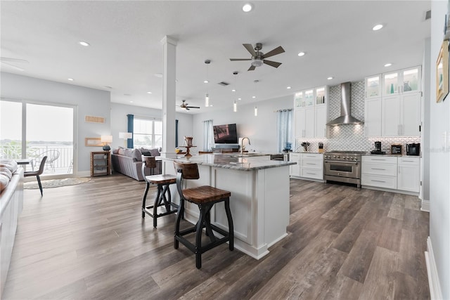 kitchen featuring wall chimney exhaust hood, ceiling fan, stainless steel stove, open floor plan, and a center island with sink