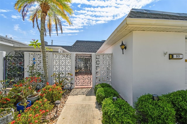 property entrance featuring a gate, stucco siding, fence, and roof with shingles