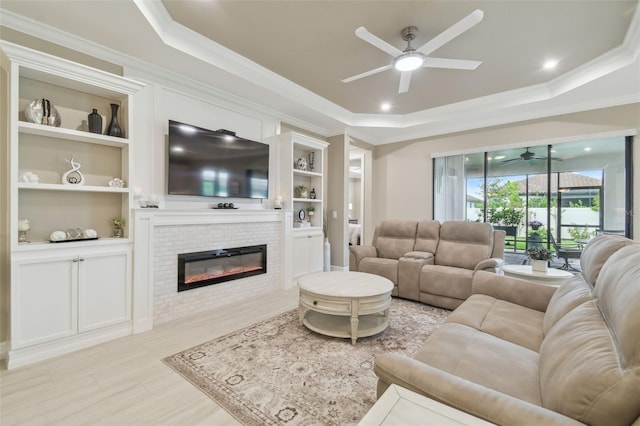living room featuring a ceiling fan, a raised ceiling, ornamental molding, a brick fireplace, and built in features