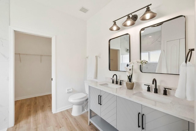bathroom featuring wood finished floors, toilet, double vanity, a walk in closet, and baseboards