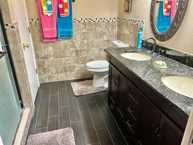 bathroom featuring tile walls, a stall shower, double vanity, toilet, and a wainscoted wall