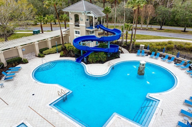 community pool with a water slide, fence, a patio area, and view of wooded area
