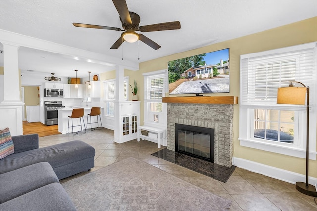 living area featuring light tile patterned floors, ornate columns, ceiling fan, baseboards, and a fireplace with flush hearth