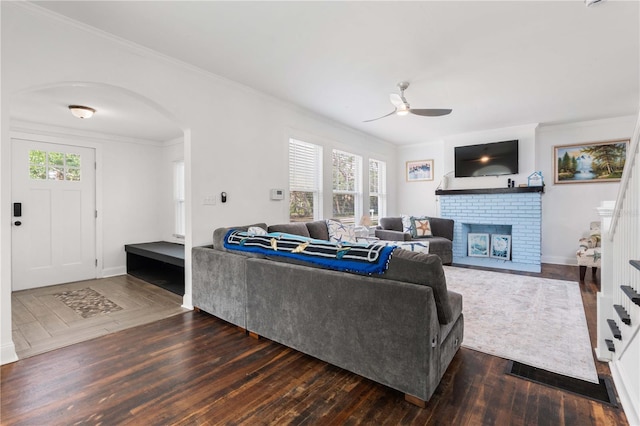 living area featuring a ceiling fan, wood-type flooring, ornamental molding, a brick fireplace, and arched walkways