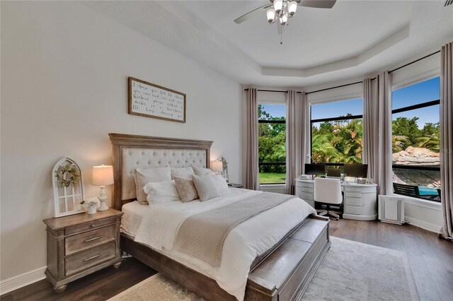 bedroom with dark wood finished floors, a tray ceiling, an office area, baseboards, and ceiling fan