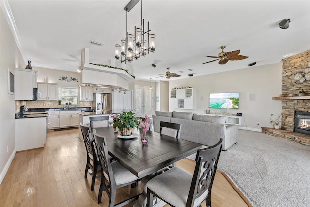 dining room with a ceiling fan, a stone fireplace, light wood finished floors, baseboards, and crown molding