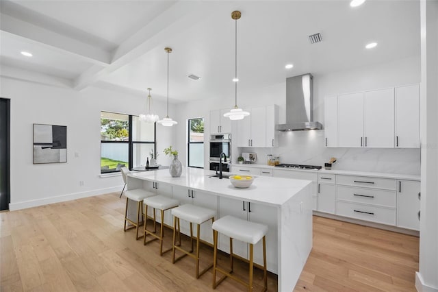 kitchen with wall chimney exhaust hood, a kitchen bar, stainless steel appliances, light wood-type flooring, and an island with sink
