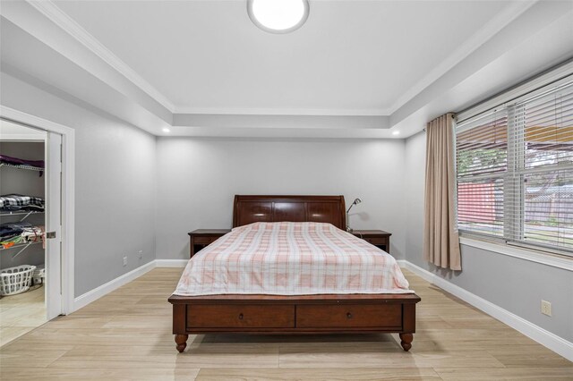 bedroom featuring light wood finished floors, a raised ceiling, baseboards, a walk in closet, and crown molding