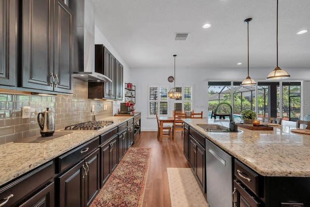 kitchen with appliances with stainless steel finishes, a sink, light wood finished floors, wall chimney exhaust hood, and backsplash