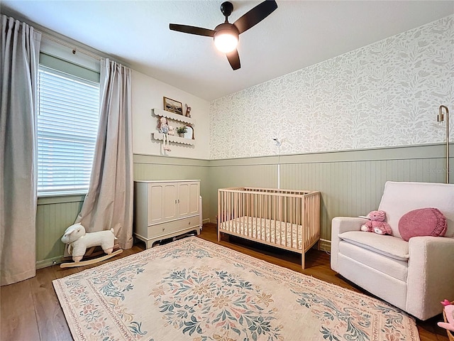 bedroom featuring a wainscoted wall, wood finished floors, multiple windows, ceiling fan, and a nursery area