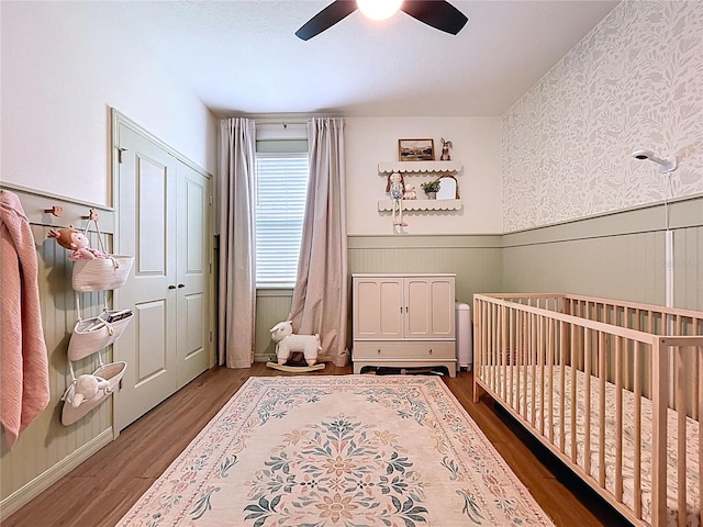 bedroom with a wainscoted wall, a crib, dark wood-type flooring, and a ceiling fan