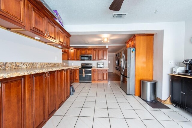 8305 75th Pl, Seminole, FL 33777 - kitchen featuring appliances with stainless steel finishes, ceiling fan, brown cabinetry, light tile patterned flooring, and light stone counters