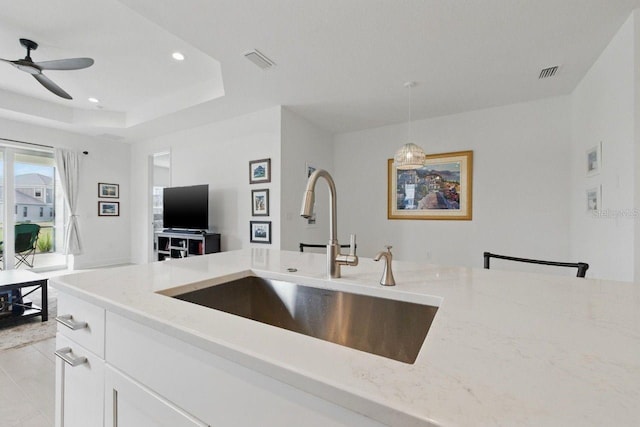 kitchen with light stone countertops, a sink, ceiling fan, hanging light fixtures, and a tray ceiling