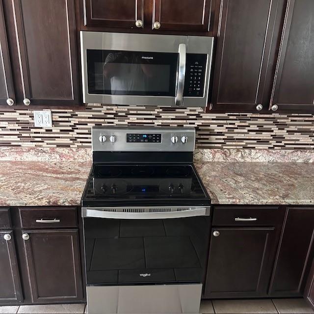 kitchen with appliances with stainless steel finishes, backsplash, light stone countertops, dark brown cabinets, and light tile patterned flooring