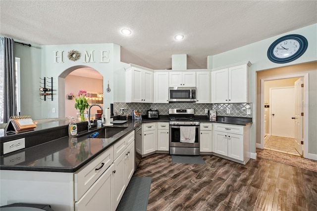 kitchen with appliances with stainless steel finishes, a sink, dark wood finished floors, dark countertops, and arched walkways
