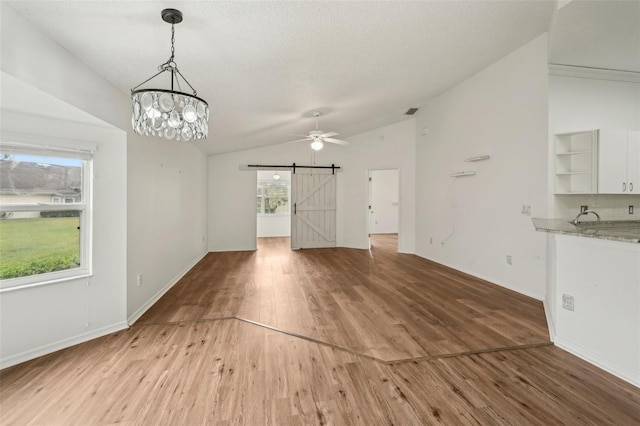unfurnished living room with a barn door, vaulted ceiling, light wood-type flooring, a ceiling fan, and a chandelier