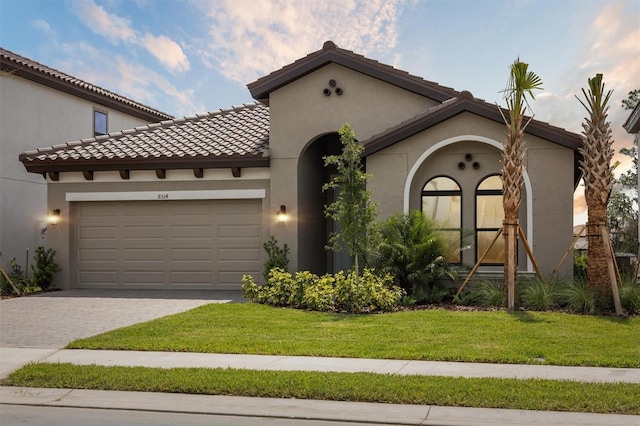 mediterranean / spanish-style home with decorative driveway, an attached garage, a front yard, stucco siding, and a tiled roof