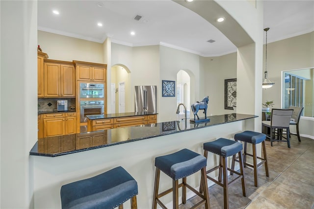 kitchen with arched walkways, appliances with stainless steel finishes, ornamental molding, a kitchen bar, and dark stone countertops