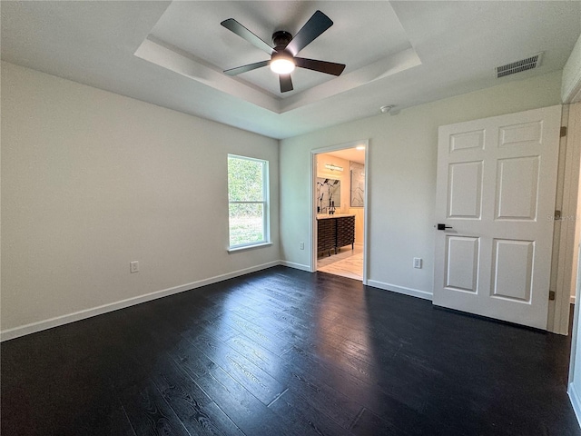 unfurnished bedroom with a tray ceiling, baseboards, dark wood-style flooring, connected bathroom, and a ceiling fan