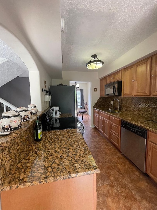 kitchen with stainless steel appliances, a sink, a textured ceiling, tasteful backsplash, and dark stone counters