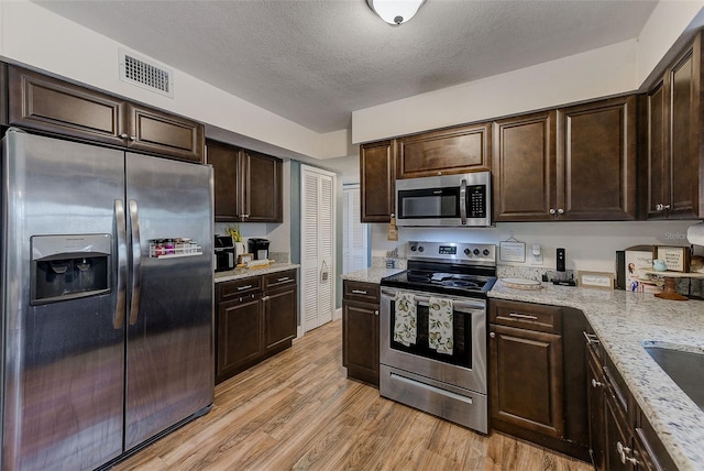 kitchen with appliances with stainless steel finishes, dark brown cabinetry, light wood-style floors, a textured ceiling, and light stone counters