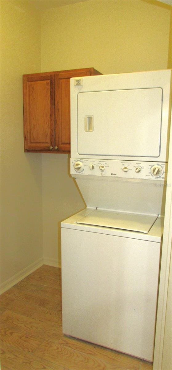 clothes washing area featuring stacked washer / drying machine, cabinet space, light wood-type flooring, and baseboards
