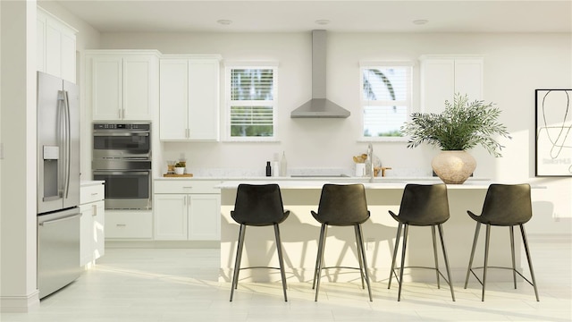 kitchen with wall chimney range hood, stainless steel appliances, a center island with sink, light countertops, and white cabinets