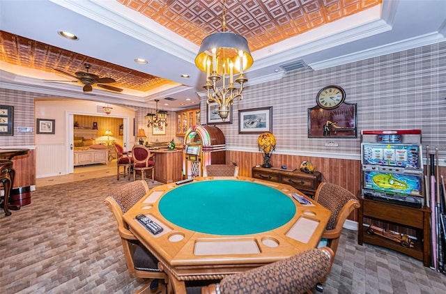 rec room with a tray ceiling, crown molding, a ceiling fan, a chandelier, and wainscoting