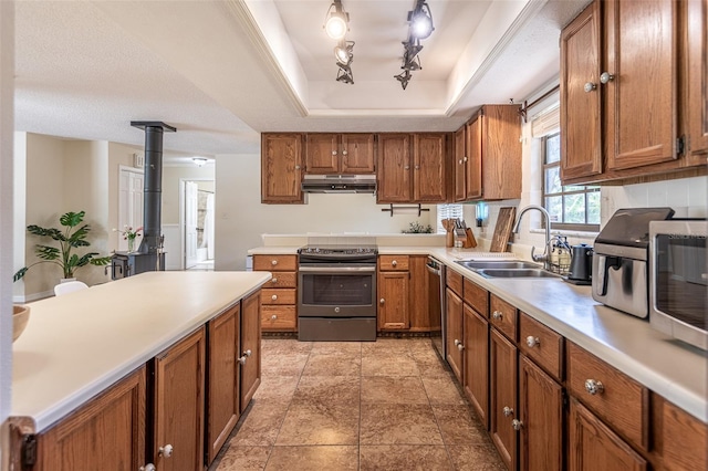 kitchen with stainless steel appliances, a sink, under cabinet range hood, a tray ceiling, and brown cabinetry