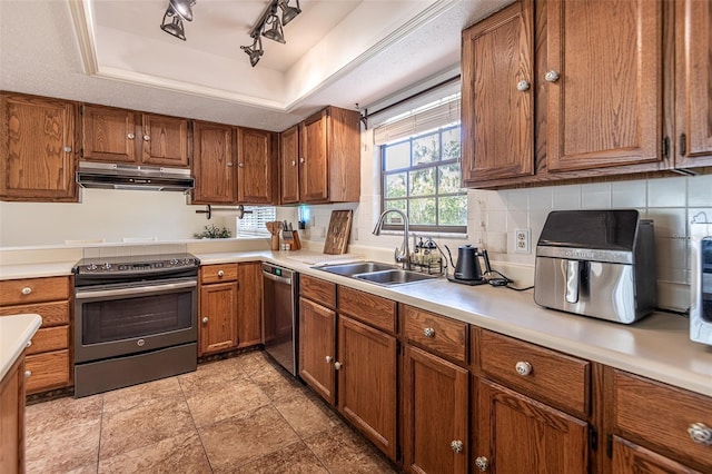 kitchen featuring appliances with stainless steel finishes, under cabinet range hood, a tray ceiling, a sink, and brown cabinetry