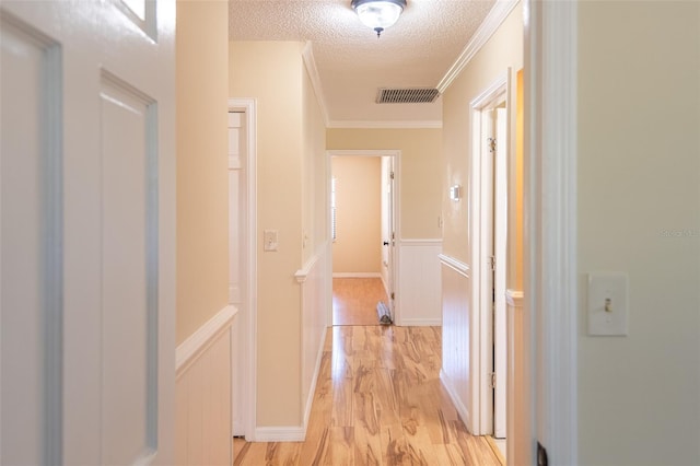 corridor featuring light wood-type flooring, a textured ceiling, crown molding, wainscoting, and a decorative wall