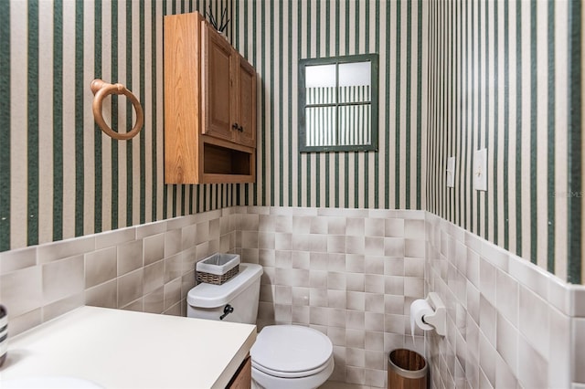 half bath with wallpapered walls, toilet, tile walls, a wainscoted wall, and vanity