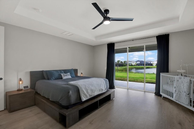 bedroom with a raised ceiling, access to exterior, wood finished floors, a water view, and ceiling fan