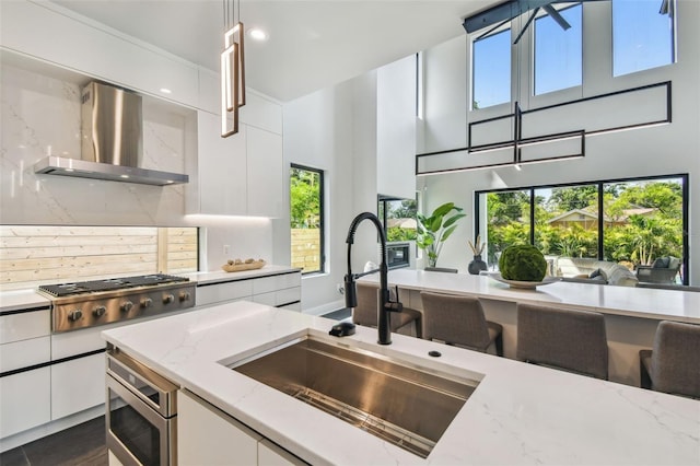 kitchen with a sink, wall chimney range hood, white cabinets, healthy amount of natural light, and a high ceiling