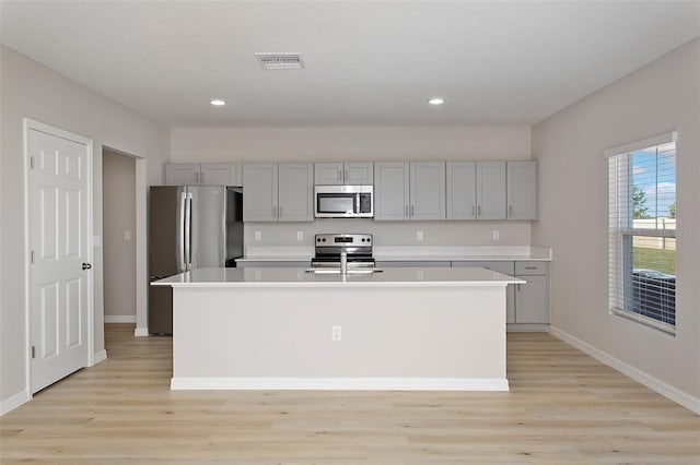 kitchen with stainless steel appliances, gray cabinets, light countertops, light wood finished floors, and a kitchen island with sink
