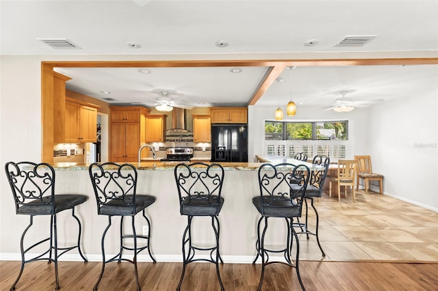 kitchen with black fridge, a peninsula, a ceiling fan, wall chimney range hood, and electric range