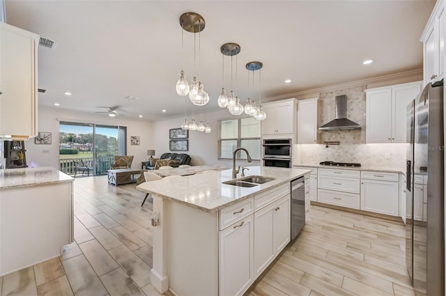 kitchen with a sink, wall chimney range hood, appliances with stainless steel finishes, open floor plan, and ceiling fan