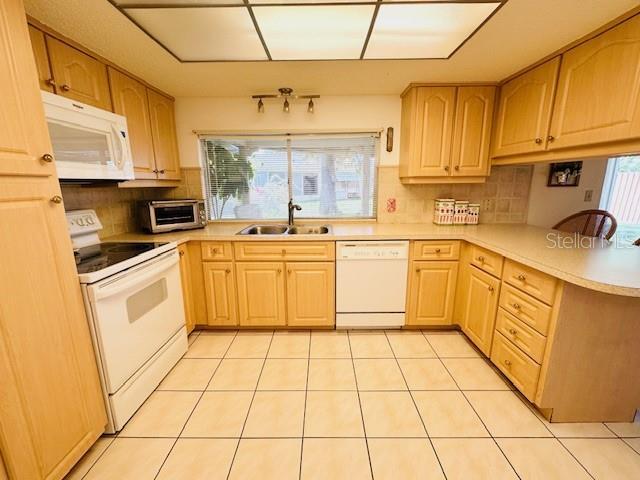 kitchen with white appliances, a sink, a peninsula, light brown cabinetry, and backsplash