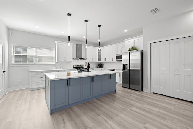 kitchen with appliances with stainless steel finishes, wall chimney range hood, white cabinetry, light wood-style flooring, and light countertops