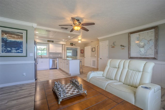 living area featuring a ceiling fan, light wood finished floors, ornamental molding, wainscoting, and a textured ceiling