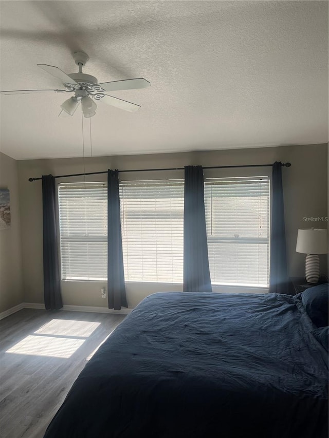 bedroom with wood finished floors, multiple windows, baseboards, a ceiling fan, and a textured ceiling