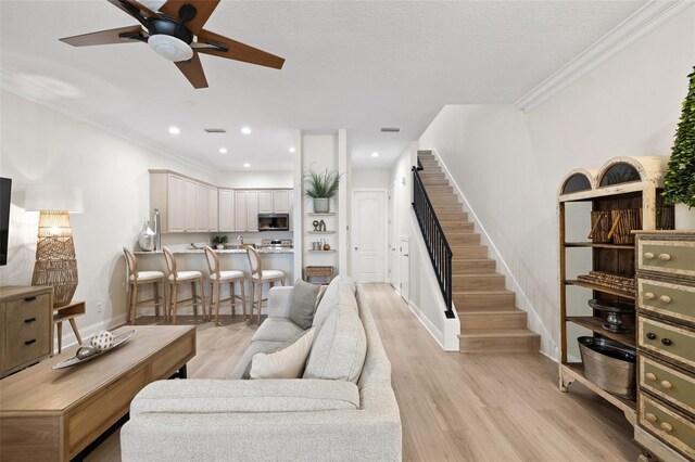 living room featuring stairs, light wood-style flooring, baseboards, a ceiling fan, and crown molding