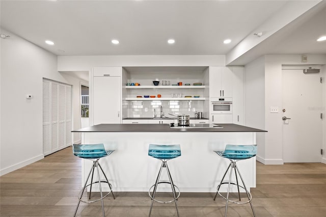 kitchen with white oven, open shelves, white cabinetry, dark countertops, and a kitchen bar